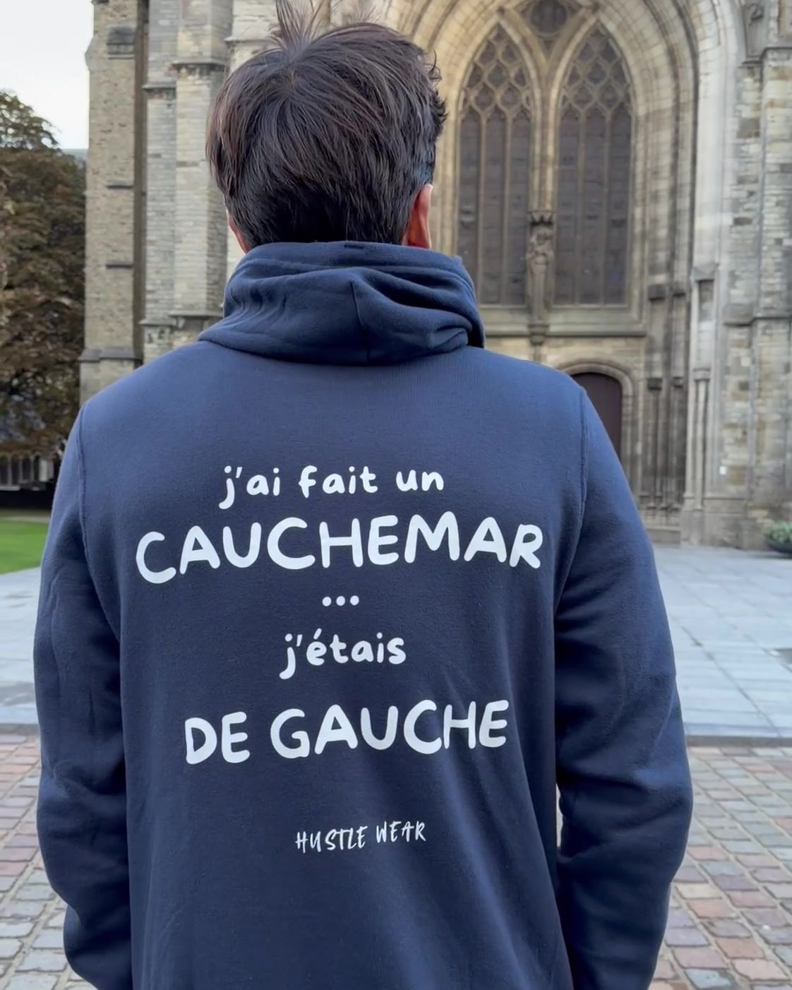 Homme de dos devant la cathédrale d'Ypres portant le pull à capuche J'ai fait un cauchemar j'étais de gauche Hustle Wear, coloris marine
