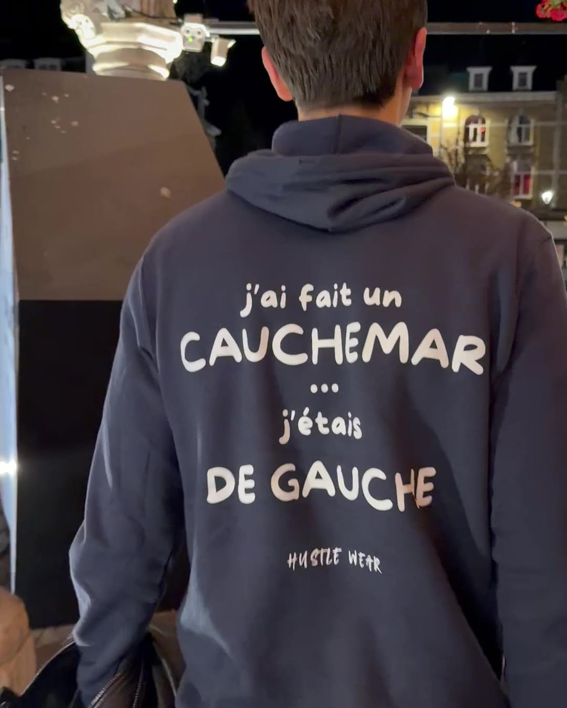 Homme de dos de nuit sur la Grand-Place d'Ypres portant le pull à capuche J'ai fait un cauchemar j'étais de gauche Hustle Wear, coloris marine