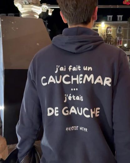 Homme de dos de nuit sur la Grand-Place d'Ypres portant le pull à capuche J'ai fait un cauchemar j'étais de gauche Hustle Wear, coloris marine