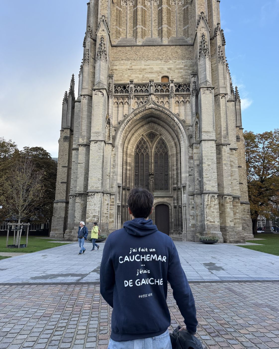 Homme de dos sur le parvis de la cathédrale d'Ypres portant le pull à capuche J'ai fait un cauchemar j'étais de gauche Hustle Wear, coloris marine