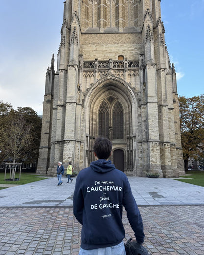 Homme de dos sur le parvis de la cathédrale d'Ypres portant le pull à capuche J'ai fait un cauchemar j'étais de gauche Hustle Wear, coloris marine