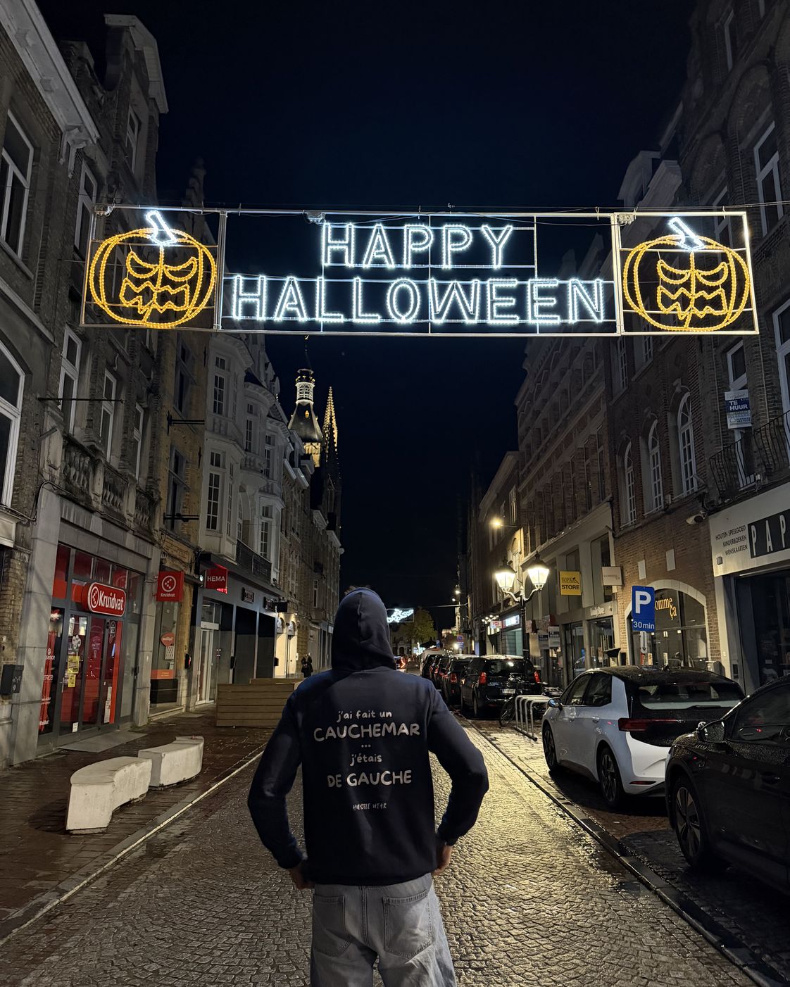 Homme de dos de nuit sous une guirlande Happy Halloween portant le pull à capuche J'ai fait un cauchemar j'étais de gauche Hustle Wear, coloris marine
