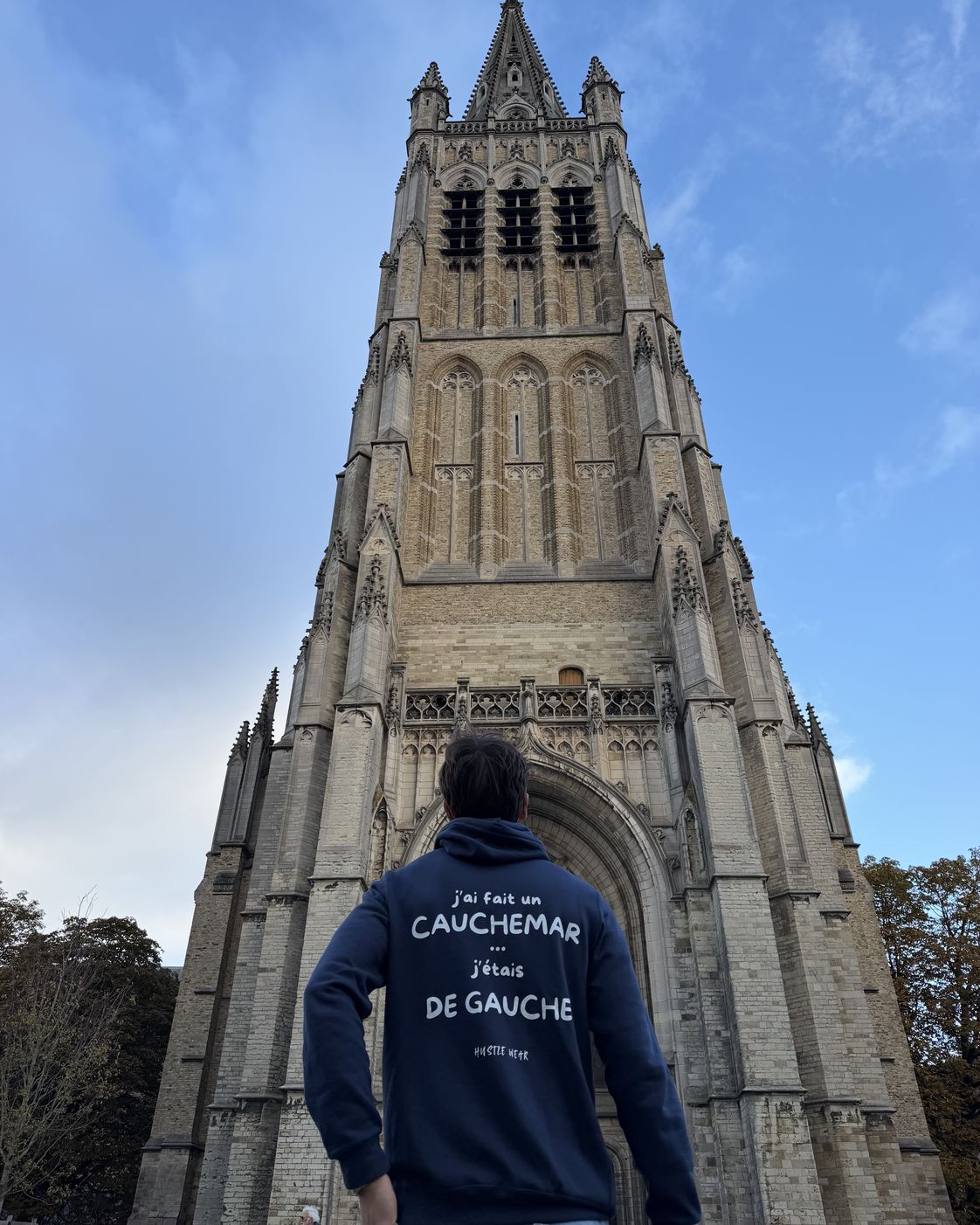 Homme de dos au pied de la tour de la cathédrale d'Ypres portant le pull à capuche J'ai fait un cauchemar j'étais de gauche Hustle Wear, coloris marine