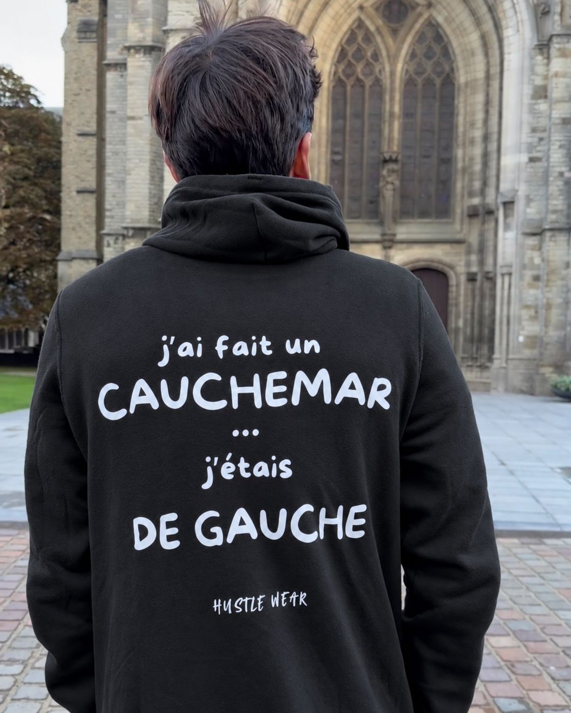 Homme de dos devant la cathédrale d'Ypres portant le pull à capuche J'ai fait un cauchemar j'étais de gauche Hustle Wear, coloris noir