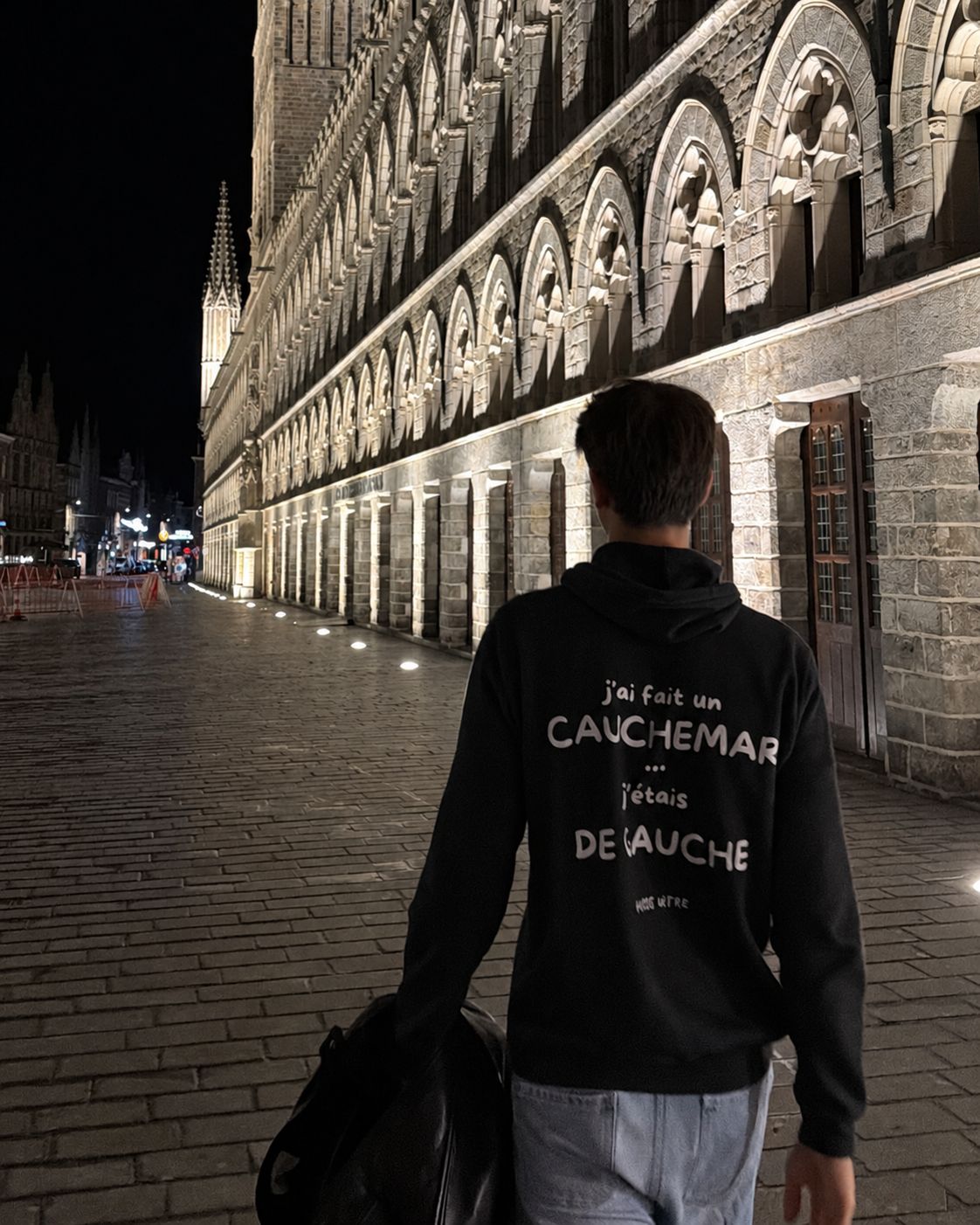 Homme de dos de nuit le long de la Halle aux draps d'Ypres portant le pull à capuche J'ai fait un cauchemar j'étais de gauche Hustle Wear, coloris noir
