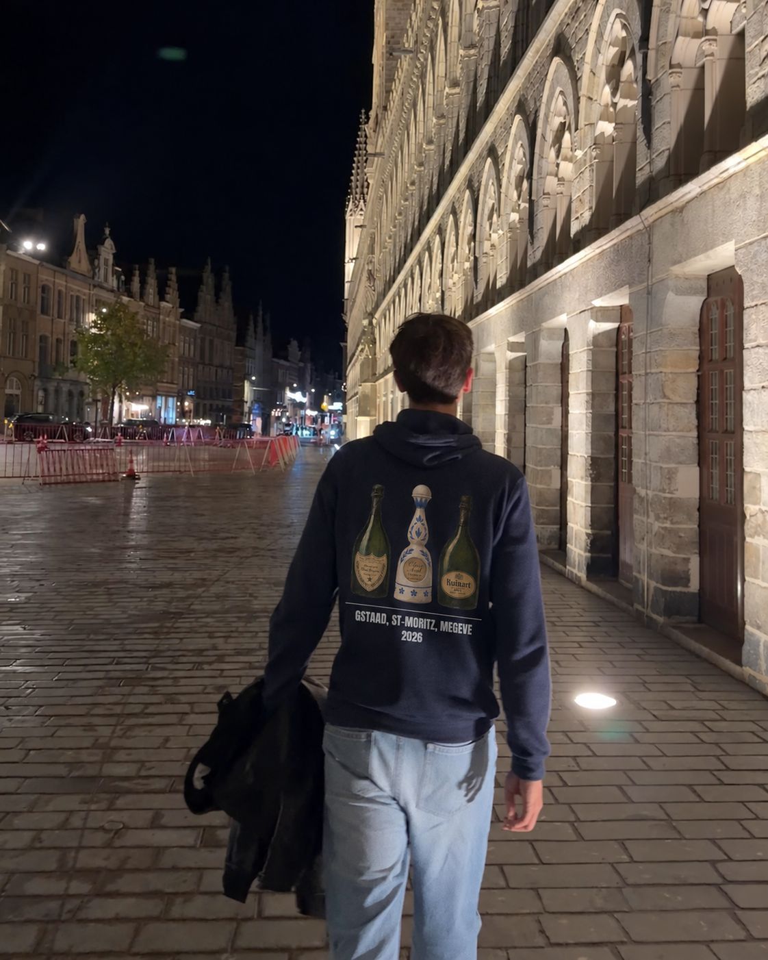 Homme de dos de nuit le long de la Halle aux draps d'Ypres portant le pull à capuche Gstaad St-Moritz Megève Hustle Wear, coloris marine