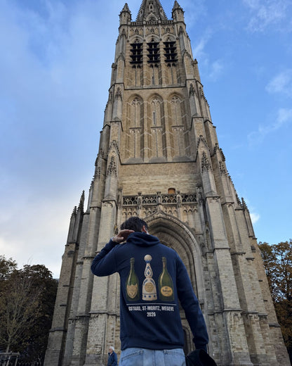 Homme de dos au pied de la tour de la cathédrale d'Ypres portant le pull à capuche Gstaad St-Moritz Megève Hustle Wear, coloris marine