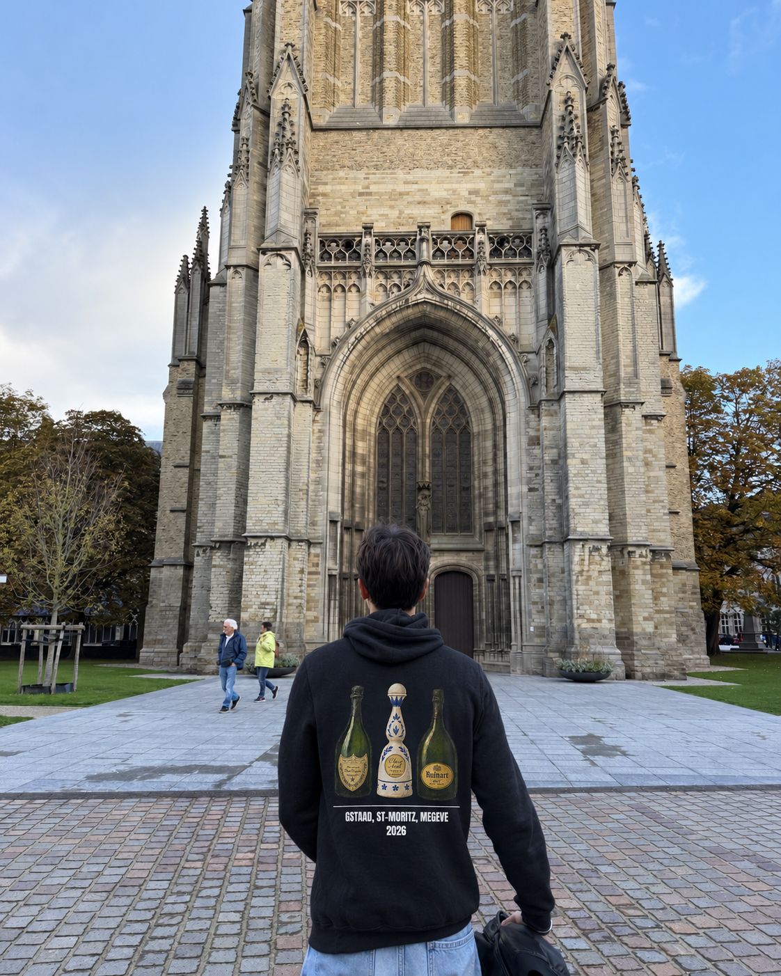Homme de dos sur le parvis de la cathédrale d'Ypres portant le pull à capuche Gstaad St-Moritz Megève Hustle Wear, coloris noir
